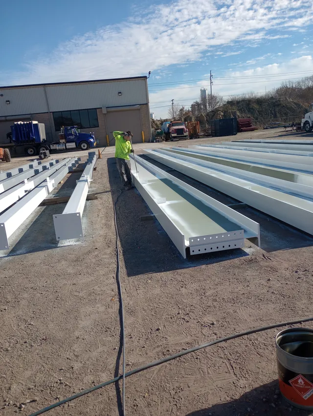 Rows of structural steel beams in a yard after prep, with crew member spraying coating